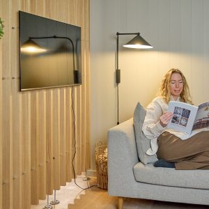 Kobieta czyta magazyn na szarej sofie obok ściany wykończonej panelami lamelowymi WoodUpp w naturalnym odcieniu dębu; na panelach zamontowany telewizor, a lamele osadzone na chromowanych wspornikach odsłaniają przestrzeń podłogi. Obok czarna stożkowa lampa ścienna oświetla kącik, tworząc eleganckie, akustyczne tło wnętrza.
