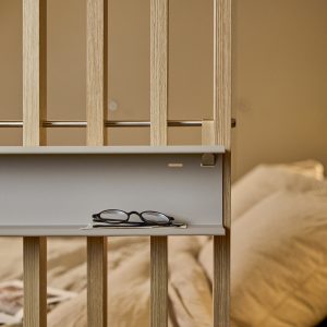 Zdjęcie przedstawia modułową ściankę WoodUpp z pionowymi, jasnymi dębowymi listwami; do niej zamocowana jest matowoszara, wąska półka-przegroda WoodUpp na metalowym uchwycie z miedzianym wykończeniem oraz cienki stalowy drążek. Na półce leżą okulary; w tle rozmyte beżowe pościele sypialni.