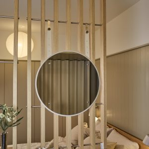 Zdjęcie przedstawia produkt WoodUpp: pionową przegrodę z jasnych dębowych listew montowanych na metalowych prętach oraz okrągłe lustro WoodUpp z cienką białą ramą, zawieszone na regulowanych jasnych skórzanych paskach, odbijające zasłonę i część przytulnej sypialni. Nowoczesne, minimalistyczne wykończenie z subtelnymi chromowanymi mocowaniami i widocznymi śrubkami.