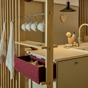 Zdjęcie prezentuje kuchenny system organizacyjny WoodUpp — dębową, wolnostojącą ramę z jasną półką na drobiazgi, na której leżą małe nożyczki, oraz stalowym relingiem, na którym wiszą białe, ceramiczne filiżanki. Widoczny jest też bordowy, filcowy pojemnik-szuflada oraz metalowe haczyki z torbami i ściereczkami.