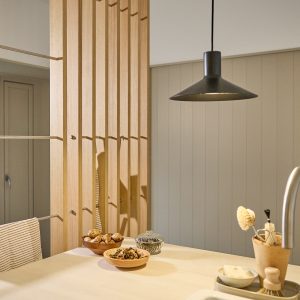 Ażurowy panel WoodUpp z pionowych, jasnych dębowych listew, zamontowany jako dekoracyjna ścianka przy blacie kuchennym; w listewach widoczne metalowe piny i cienkie poziome pręty, obok lampa wisząca i drewniane miski z orzechami, tworzące naturalny, minimalistyczny wystrój z naturalnym wykończeniem i subtelnymi cieniami.