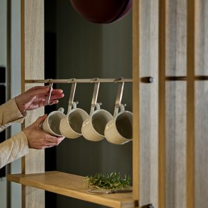 Zdjęcie przedstawia regał kuchenny WoodUpp z jasnego drewna z zamocowanym metalowym relingiem i pięcioma metalowymi hakami WoodUpp, na których wiszą ceramiczne, kremowe kubki; w górnej części widać ręce zawieszające kubek, na półce gałązka ziół o praktycznym, minimalistycznym wykończeniu i kubkach w matowym szkliwie.