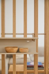 Zdjęcie prezentuje produkt WoodUpp: ażurową ściankę z pionowych dębowych listew z metalowym prętem i zamocowaną beżową półką. Na półce znajdują się dwie drewniane miseczki WoodUpp — jedna większa o naturalnym rysunku słoi, druga mniejsza z wyraźnym wzorem słojów i minimalistycznym skandynawskim designem.