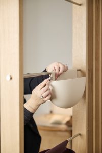 Ręce regulują beżowy, półokrągły pojemnik na relingu modułowego systemu WoodUpp: metalowa, stalowa listwa i plastikowy uchwyt mocujący. Zdjęcie pokazuje jasne, dębowe panele garderoby, metalowy pręt i dolny wieszaczek z ciemnym pokrowcem — praktyczne akcesoria WoodUpp do przechowywania, idealne do małych przestrzeni i organizacji.