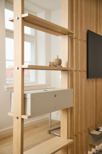 Zdjęcie przedstawia elegancką ażurową ściankę-półkę WoodUpp z jasnego dębowego drewna o otwartej, minimalistycznej konstrukcji, łączoną stalowymi śrubami, z zamontowaną metalową beżową skrzynką na kable WoodUpp oraz dekoracyjną szpulą sznurka; w tle widać drewniane lamele i fragment telewizora oraz naturalne światło wpadające z okna.